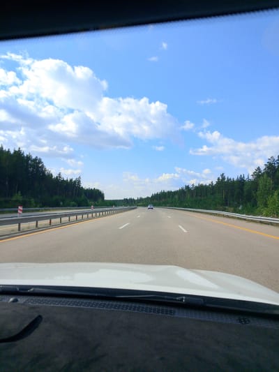A first-person view from inside a vehicle traveling on a highway. The road ahead is paved and has clear lane markings, with a yellow line along the right edge and white lines dividing lanes. A metal guardrail runs along the left side of the road, bordered by red and white reflective markers. To the left of the guardrail is a dense line of dark green coniferous trees. The sky is bright blue with scattered white clouds. Further down the road, two white cars are visible. The foreground shows the interior of the vehicle, including the white hood and a dark dashboard. The image appears to be taken in a rural or semi-rural area, possibly in the vicinity of Алтынды ауылдық округі, Kazakhstan, given the presence of a highway and forested areas.