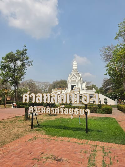 The image shows a white temple building in Historic City of Ayutthaya, Thailand. A sign in Thai characters stands in front of the temple. The surrounding environment consists of trees, grass, and a brick path. The sky is blue with some clouds.