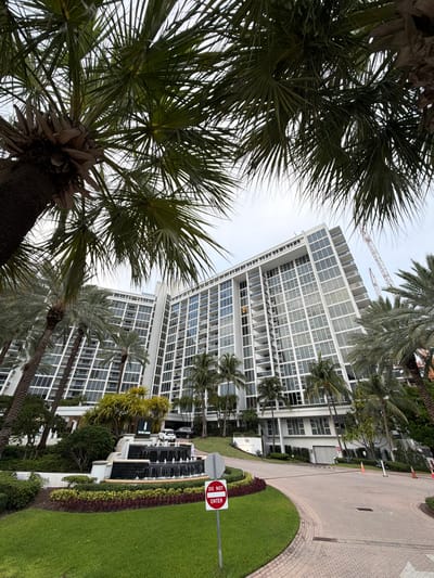 The image captures a frontal view of the Ritz-Carlton Bal Harbour, a luxury hotel located at 10295 Collins Avenue in Bal Harbour Village, Florida, USA. The building is a tall, modern white structure with numerous glass-paneled balconies and windows. In the foreground, palm trees with lush green fronds are prominently featured, some partially obscuring the view of the hotel. A circular driveway leads to the entrance, which is adorned with a multi-tiered fountain and manicured landscaping featuring green grass and colorful bushes. A "Do Not Enter" sign is visible on the right side of the driveway. Two vehicles are parked near the entrance. Construction cranes are visible in the background, suggesting ongoing development in the area. The sky is overcast, and the overall impression is one of a tropical, upscale resort.