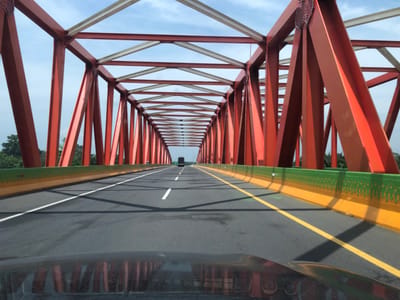 The image provides a first-person perspective from inside a vehicle driving through a large, enclosed metal truss bridge in Gohor Lama, Indonesia. The bridge's main structure is painted a striking red, composed of numerous interconnected beams and girders, with some upper cross-beams appearing white or silver. These form a repetitive, geometric pattern overhead and along the sides, creating a tunnel-like passage that converges into the distance.
The asphalt roadway has clear white solid and dashed lane markings in the center, and a solid yellow line on the right side. Distinct shadows from the bridge's framework are cast across the road surface. Along both sides of the road, prominent guardrails feature a bright green upper section with a decorative patterned cut-out, surmounting a solid yellow lower section.
In the far distance, a large, dark-colored truck is visible, driving ahead in the same direction. Beyond the bridge structure, glimpses of a partly cloudy sky and abundant green trees and foliage are visible, suggesting a natural or semi-rural setting. The bright illumination indicates it is daytime, likely late morning or early afternoon, under fair weather conditions. The foreground reveals a reflection on a vehicle's dashboard or hood, confirming the vantage point. There is no visible text in the image.