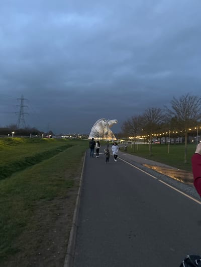 The image depicts an outdoor scene at dusk or early evening within The Helix park in Falkirk, Scotland, United Kingdom. In the middle-ground, a large, illuminated, silvery-grey equine head sculpture, identifiable as one of The Kelpies, stands prominently. Behind it, a second, less distinct horse head sculpture is partially visible. An asphalt pedestrian pathway occupies the lower half of the frame, leading towards the sculptures. Four individuals are present on this path: three adults and one child. They are walking away from the camera towards the sculptures. From left to right on the path, an adult male in dark clothing, an adult female in light clothing, a child in dark clothing, and another adult female in a white jacket are discernible. To the left of the pathway is a grassy embankment. To the right, a grassy area features trees adorned with string lights, and reflective puddles are visible on the path's edge. In the far left background, a tall metal electricity pylon stands against an overcast, dark blue-grey sky. Artificial lighting from park lamps and the illuminated sculptures provides the primary illumination for the scene. A blurred red sleeve is partially visible in the bottom right corner, suggesting a first-person perspective.
