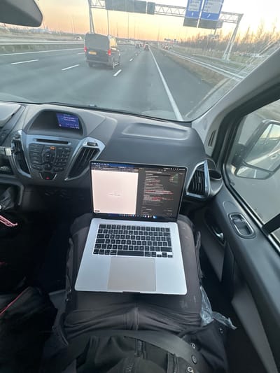 The image depicts the interior of a vehicle's front passenger area and the view through its windshield onto a multi-lane highway. A silver MacBook Pro laptop rests open on the lap of a person wearing dark pants and a dark jacket. The laptop screen is split, displaying a web browser with a loading spinner on the left side and what appears to be code or terminal output on the right.

The vehicle's dashboard is visible, featuring an infotainment system that displays "BT Audio," "PlayMode: None," and "00:04." Air vents are located on the dashboard and passenger door. A black bag and pink braided material are visible on the floor to the left of the person's legs.

The exterior scene shows a highway with multiple lanes of traffic. The sky displays an orange-yellow hue, suggesting either sunrise or sunset. In the immediate foreground, directly ahead in the same lane, is a black van with a visible Dutch license plate: TLR-90-J. A large overhead gantry sign structure spans the highway in the distance. Blue highway signs attached to this gantry clearly indicate "D-4b Verlading," "Cargo," and "Hoofddorp," confirming the location in the vicinity of Hoofddorp, Netherlands. Other partially visible text includes "Oude Meer" and "Cargo Rijk." Trees and light poles line the right side of the highway. Other vehicles are present further down the road.