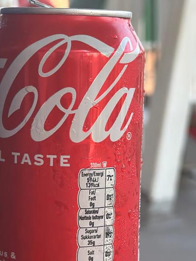 This is a close-up, angled shot of a condensation-covered can of Coca-Cola. The iconic red can features the white Coca-Cola logo and the text "REAL TASTE" in white lettering. A nutrition label is visible on the right side of the can, detailing energy content, fat, saturates, sugars, and salt. The can appears to be in an outdoor setting, possibly on a table, with a blurred background of what could be foliage or other outdoor elements. The condensation on the can suggests it is a cold beverage on a warm day. The overall impression is one of refreshment and enjoyment of a popular soft drink.