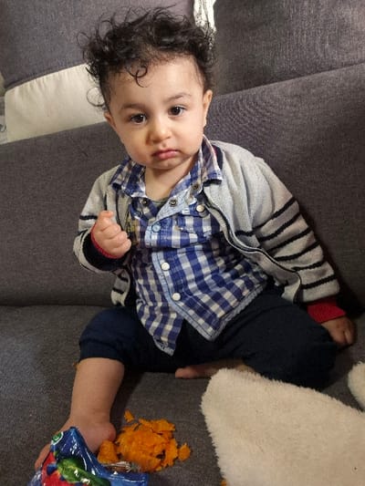 An infant is seated on a gray fabric sofa indoors in Melle, Belgium, facing directly towards the camera with a slight frown and downturned mouth. The infant has dark, curly hair and is wearing a blue and white plaid button-up shirt over a lighter undershirt, a gray and white striped cardigan with red cuffs, and dark blue pants. The infant's left hand is slightly fisted, and the right hand rests beside the body. Bare feet are visible. To the infant's left, a pile of orange, mashed food, possibly sweet potato, is scattered on the sofa, next to an open blue and red snack package. A white, fuzzy object, possibly part of a blanket or toy, is visible on the sofa to the infant's right. A white pillow is partially visible behind the infant on the sofa.