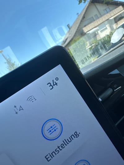 The image shows a close-up of a car's digital display, reflecting an outdoor scene with a blue sky and a building. The display shows the temperature as "34°", a Wi-Fi symbol, and the text "Einstellung.". The building appears to be a house with a peaked roof and windows. The scene is likely captured on a sunny day. The reflection on the display includes what appears to be text written on a window, which reads "32M". The car interior is visible in the lower right corner, including a gray lever. The overall context suggests a car parked in a residential area, possibly in Brackenheim, Germany, given the reflection of what might be local text or signage and the architectural style of the house.