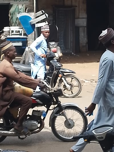 The scene captures an active street in Kofar Mazugal, Nigeria, under bright natural light. Three adult males and multiple motorcycles are present. In the left foreground, a man in a brown tunic and patterned cap sits astride a black motorcycle with spoked wheels, a yellow container affixed to the rear. His hands are on the handlebars. Centrally positioned in the midground, a man in a light blue tunic and patterned cap stands next to a black scooter with alloy wheels, looking directly towards the camera. To the right, partially visible, a third man in a light blue tunic and dark patterned cap walks forward, holding a small object in his right hand. The ground is unpaved, composed of dirt and compacted earth. The background shows parts of buildings, including a dark doorway, and indistinct market-style elements and vehicles.
