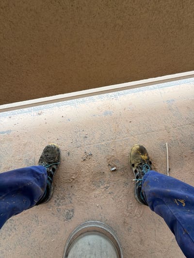 This is a first-person perspective, eye-level shot of a person wearing blue work pants and muddy work shoes. The scene appears to be outdoors, on a tiled floor covered in a light brown, dusty material, possibly soil or construction debris. In the foreground, the person's feet are visible, with their right shoe in the center-right and their left shoe towards the center-left. The shoes are dark and heavily soiled, with blue laces. The pants are a deep blue and also show signs of being covered in the same dust.

Above the tiled floor, there is a distinct horizontal line, suggesting a doorway or a transition to a different surface. Above this line, there is a textured, brown surface, possibly a wall or the underside of an overhang. The lighting suggests it is daytime.

Based on the context provided, this scene is located in the city of Příbram, Czechia. While the specific location within Příbram is not visually discernible from the image itself, it appears to be an outdoor area that is either under construction or has recently experienced some form of disturbance leading to the accumulation of dust and debris on the ground and footwear. There are no other individuals or discernable structures that would indicate a specific landmark or area within Příbram. The image captures a moment of everyday work or activity in an urban or suburban environment.