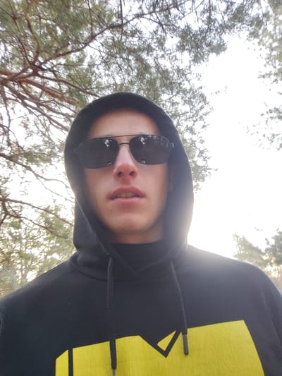 A photograph depicts a single young male individual, appearing of light complexion, positioned outdoors in Бърдарите, Bulgaria. He wears a black hooded sweatshirt with the hood covering his head and dark sunglasses obscuring his eyes. A dark, high-collared garment is visible beneath the sweatshirt. A yellow geometric graphic, resembling a partial 'M', is visible on the front lower portion of the black sweatshirt. The individual's mouth is slightly ajar. The background consists of out-of-focus evergreen tree foliage against a bright, possibly overcast or backlit sky. The individual appears to be posing for the camera, which is positioned below eye level.