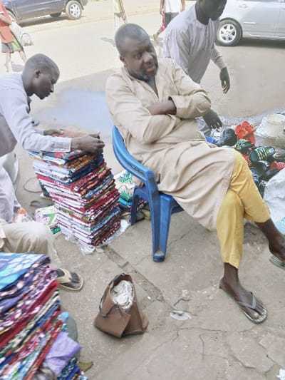 The scene is an outdoor market area in the city of Kungulu School, Zambia. In the foreground, a dark-skinned man with a beard, wearing a light-colored tunic and yellow trousers, sits on a blue plastic chair with his arms crossed, looking towards the left. He wears dark flip-flops. To his immediate left, another dark-skinned man in a light grey long-sleeved shirt is bent over, actively arranging a tall stack of colorful, folded textile fabrics on the ground. A second, partial stack of similar fabrics is visible further left. To the right of the seated man, assorted shoes and sandals are displayed on the ground, accompanied by white containers. A small, open brown bag with white contents rests on the cracked concrete ground. In the background, a third dark-skinned man in a light-colored garment is partially visible behind the seated individual. A paved road is present in the mid-ground, featuring a dark SUV, a light-colored sedan, and several blurred figures walking or standing. The ground is an uneven, worn concrete or tarmac surface.