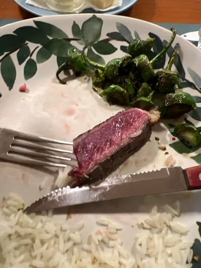 A plate holds a partially consumed meal. A slice of rare steak, skewered on a fork, rests beside a serrated knife. Pimientos de Padrón are clustered on the left, their dark green color contrasting with the white plate. A mound of cooked rice occupies the right side. The plate itself has a leaf pattern. A small blue plate with food is partially visible on the left.
