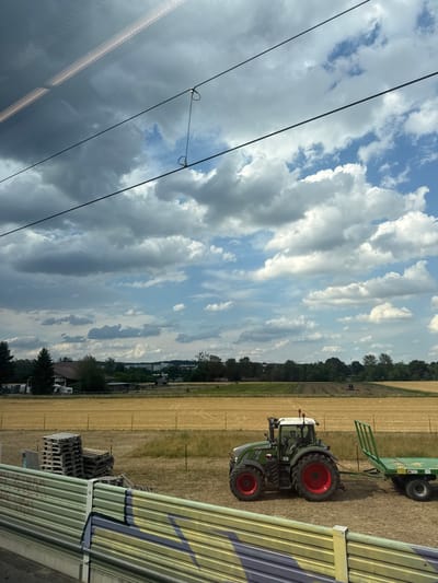 This is a media file showing a scene in Renningen, Germany. The main subjects are a green tractor with red wheels and a green trailer it is pulling, parked on a dry, golden field. The field appears to be recently harvested. In the background, there are trees and some buildings, suggesting a rural or semi-rural area. There are also some power lines visible in the foreground, running diagonally across the frame. The sky is blue with dramatic, puffy white and grey clouds, indicating a mix of sun and clouds, possibly late afternoon.
The tractor is a Fendt model, with "FENDT 724" visible on its side. The trailer has a ramp-like structure at its rear. To the right of the field, there is a section of a structure with yellow and green corrugated metal siding, and some stacked wooden pallets. The overall impression is of agricultural work, likely a pause in operations due to the weather or time of day. There are no people clearly visible in the image. The scene conveys a sense of calm and quiet, with the vast sky and the stationary machinery. There are no discernible emotions conveyed, but the overall mood is peaceful. No specific event is explicitly depicted, other than the presence of farming equipment in a field.