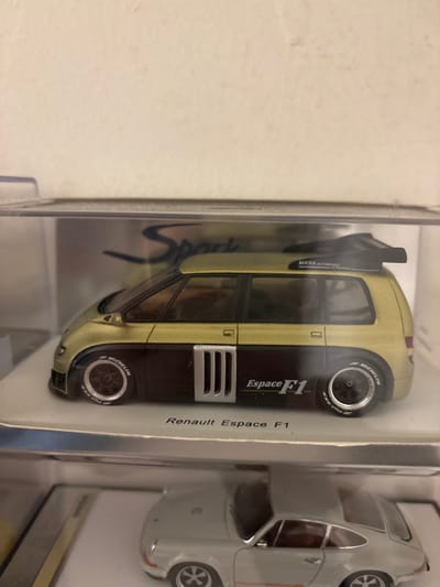 The image displays a close-up, eye-level view of two miniature model cars stacked vertically within clear plastic display cases against a plain, light-colored background.

The upper display case contains a 1:43 scale model of a Renault Espace F1. The model is two-toned: the upper section, including the roof and hood, is a metallic light gold or bronze, while the lower body is dark brown or black. It features a prominent black rear spoiler marked "MATRA AUTOMOBILE," silver vertical intake vents on the lower side panel, and "Espace F1" text in gold and black on the dark body section. The wheels are black with polished rims and "MICHELIN" branding clearly visible in white on the tire sidewalls. The model rests on a white base with "Renault Espace F1" printed in black text. A 'Spark' logo is partially visible on the silver backing card behind the model.

The lower display case, partially visible, contains a model of a light grey or silver sports car, identifiable as a Porsche 911 variant due to its distinct roofline and rear engine cover detail. The model exhibits a multi-spoke wheel design.

The environment appears to be an indoor setting, likely a domestic or hobbyist display area, with a smooth, off-white wall serving as the background. No individuals are present. The scene is static.