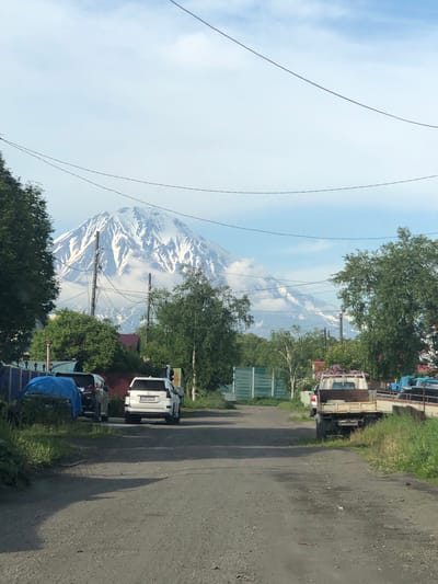 The image shows a dirt road in a semi-rural area with a large, snow-capped mountain in the background, likely a volcano. The road is flanked by sparse vegetation, trees, and a few buildings with fences. Several vehicles are parked along the side of the road, including a white SUV, a pickup truck, and another vehicle covered by a blue tarp. Power lines are visible crisscrossing the sky. The sky is partly cloudy with a blue hue. The overall scene suggests a remote location, possibly on the Kamchatka Peninsula, given the prominent volcanic landscape.