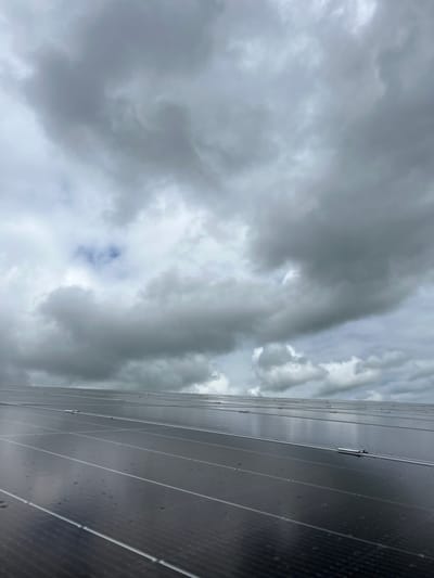 The image shows an overcast sky over Grange Ville, Ireland, with dense grey clouds filling most of the frame. The bottom right corner of the image features an array of wet solar panels angled upward, indicating they are likely mounted on a roof or ground structure. Raindrops are visible on the solar panels.