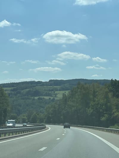 This is a landscape photograph taken from a moving vehicle on a highway. The road curves to the right, with guardrails on both sides. There are several vehicles on the road, including a white RV on the left and a dark-colored sedan ahead in the right lane. The environment is a hilly, forested area under a blue sky with scattered white clouds. The scene appears to be in Malmedy, Belgium, given the terrain and vegetation consistent with the Ardennes region.
