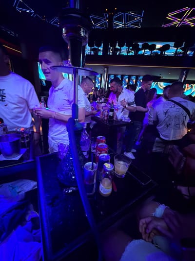 A group of people are gathered around a table in a dimly lit nightclub. The atmosphere is vibrant, with blue and purple lights illuminating the scene. A hookah pipe is prominently displayed on the table, with smoke possibly rising from it. Several drinks are scattered around, including cans of Coca-Cola and a bottle of what appears to be juice. The individuals in the image are dressed casually, suggesting a relaxed evening out. The nightclub setting, with its modern decor and lively ambiance, points to a social gathering or a night of entertainment in Nesebar, Bulgaria.
