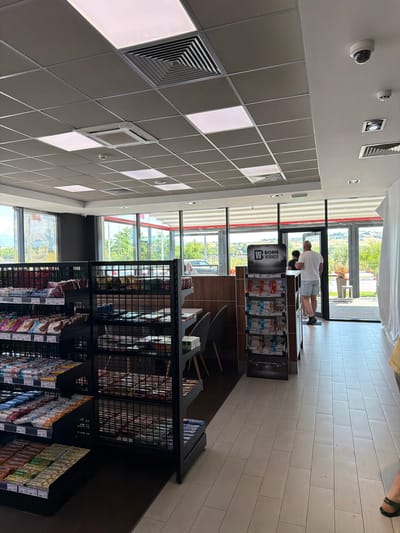 The image shows the interior of a convenience store, likely a gas station mini-mart, in Chuchuligovo, Bulgaria. The store features a drop ceiling with square white light panels and a ventilation system. Merchandise is displayed on black metal shelving units. In the foreground, shelves are stocked with various food items, including candy bars and packaged snacks. In the background, a man in a white t-shirt and blue shorts stands near the exit, looking towards the outside. Another person is partially visible behind him. The exit leads to an outdoor area with greenery and a visible road. A display stand with "BORN WINNER" branding is positioned near the entrance. The floor is tiled with light-colored rectangular tiles. A security camera is visible on the ceiling.