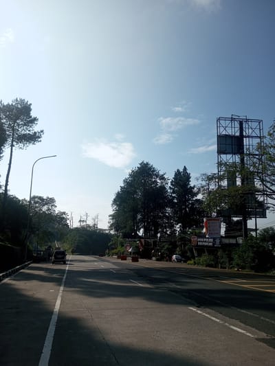 The image shows a wide, paved road in a mountainous area, likely in a tropical region. There are trees on either side of the road, and a large billboard is visible on the right side. A building with a sign that reads "POS POLISI PIR PUNCAK" is visible near the billboard. A few vehicles are present on the road. The sky is clear and blue. There is no specific indication of Phouviang-Gnai, Laos.