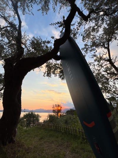 An outdoor, low-angle, full shot depicts a dark gray paddleboard leaning against a thick, dark tree trunk. The paddleboard is positioned diagonally in the frame, with its top end obscured by foliage. The paddleboard has orange markings on its side and text that reads "ITIWIT." The tree branches are silhouetted against a sky filled with clouds tinged with orange, pink, and yellow hues, indicating a sunset. In the background, a body of water stretches to the horizon, with distant mountains visible. A low wooden fence and green grass are in the foreground. The image appears to be taken in a natural setting, possibly near a lake or the sea. The overall mood is serene and tranquil. The location is not specified in the image or the prompt, but the prompt asks to include details about "the city of Garda, Italy". Without further information from the image, the city of Garda cannot be identified.