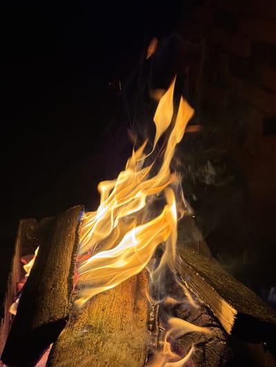 The image shows a close-up of a campfire at night, with flames and burning logs. The background is dark, with a hint of a brick structure on the right side. There are no people visible in the image. The fire is actively burning, with vibrant orange and yellow flames licking upwards. Several pieces of wood are visible, some appearing charred and others still catching fire. Wisps of smoke are also present, contributing to the atmosphere. The lighting is primarily from the fire itself, casting a warm glow on the wood and creating dramatic shadows. The overall impression is one of a cozy and natural setting, likely outdoors. It is not possible to determine the exact location within the city of Maksimovo, Russia, from the image alone, as it does not contain any recognizable landmarks or identifying features of the city.