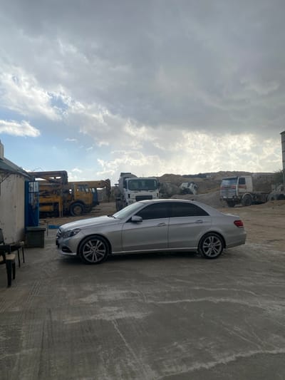 The scene depicts an outdoor industrial or construction yard in Dahiyat Harasta, Syria, under a partly cloudy sky. In the foreground, a grey, uneven concrete or dirt surface extends. A silver Mercedes-Benz E-Class sedan is parked facing left in the midground, its driver's side visible. To the far left, partially visible, are two dark chairs and a light-colored building with a blue entrance, adjacent to a green container. In the background, an array of heavy machinery is present: a yellow crane truck is positioned on the left, a white concrete mixer truck is visible behind the Mercedes, and an older white and red truck chassis sits further to the right. Piles of earth, gravel, and construction debris characterize the distant background, with a partially obscured or overturned vehicle visible within them. A tall, grey cylindrical structure rises on the far right. No individuals are visible in the frame.
