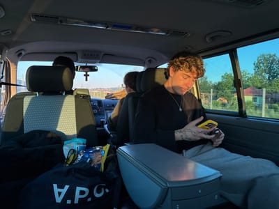 The interior of a van is captured, with three individuals visible. The primary subject, a young man with curly hair and tattoos on his hands, is seated in the back, looking at a yellow smartphone. He is wearing a black long-sleeved shirt and gray sweatpants. To his left, a gray storage console is visible.
In the front seats, the back of the driver and passenger's heads are visible. A rearview mirror with a cross hanging from it is above them.
A bag with the letters "V.P.C." on it is in the foreground, alongside a snack and eyeglasses. The window to the right reveals an outdoor scene with trees and a few houses, likely during the day due to the light.
Based on the prompt, the location is in the city of Loznica, Serbia.