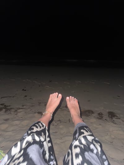 A pair of bare feet with white painted toenails are visible in the foreground, resting on a sandy beach. The feet are adorned with a colorful beaded anklet on the left ankle. The person is wearing patterned pants with a black and white design. The background depicts a dark, sandy beach with scattered seaweed and faint footprints, extending towards the dark expanse of the ocean and the night sky. The location is the city of Mare Anglaise, Seychelles.