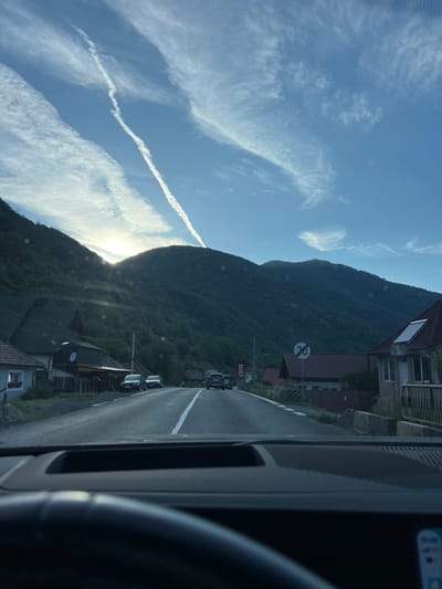 This is a roadside view from a vehicle dashboard in Robești, Romania. The scene depicts a two-lane asphalt road curving through a village nestled at the base of a forested mountain range. The sun is setting, casting a warm glow and long shadows. A prominent contrail stretches across the bright blue sky, intersected by wispy clouds. Several houses with red-tiled roofs line the road, with cars parked in front of some. A speed limit sign indicating "30" is visible on the right side of the road. The perspective is from inside the car, looking out through the windshield, with the upper portion of the dashboard visible in the foreground.