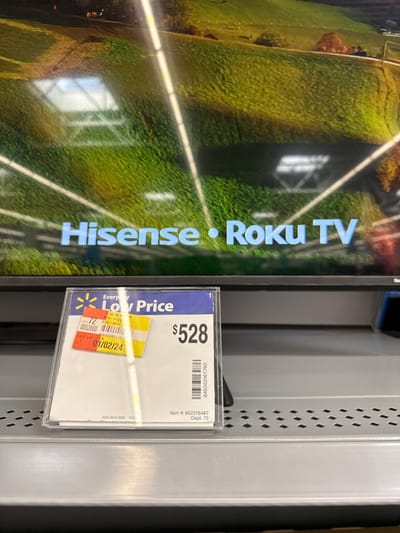 An indoor, close-up shot captures a television on display in a retail setting, likely a Walmart, given the visible branding. The screen of the television, which is rotated counter-clockwise, shows an outdoor scene of a large, lush green field with white lines, possibly a sports arena or a park. Prominently displayed on the TV screen is the text "Hisense • Roku TV" in a light blue font, indicating the brand and smart TV platform.

To the right of the TV, a clear plastic holder attached to a shelf displays a price tag. The most prominent detail on the tag is a large "$528" indicating the price. Above the price, the text "Everyday Low Price" is visible, along with the Walmart logo. Below the price, a date "01/02/24" is printed, possibly indicating a pricing update or expiration. Other information on the tag includes "Item #: 652316487," "Dept. 72," and a barcode with the number "846042061760." Additional smaller numbers and codes are present, such as "3-2186," "0052800," "120325-0012-," "CASE QTY A," and "4604206176." The background features a gray perforated metal shelf. No people are visible in the image.