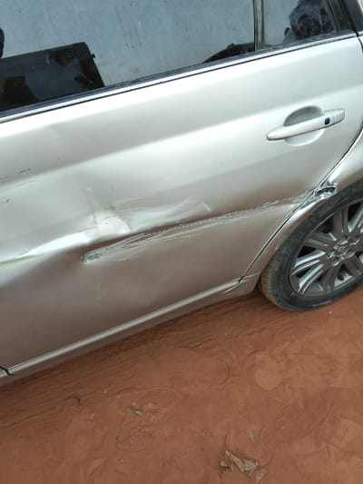 A damaged, tan-colored car shows significant dents and scrapes along the rear passenger side door. The damage extends from the middle of the door, near the door handle, back toward the rear wheel. The tire and wheel well also appear to have been impacted. The car is parked on red dirt or soil. The window of the car is visible, reflecting a dark object.