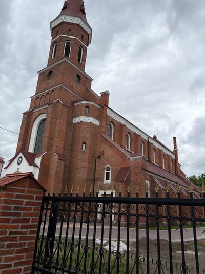 This is a low-angle, full shot of the Holy Resurrection Cathedral in Kolomyia, Ukraine, captured on an overcast day. The red brick cathedral is the primary subject, with its tall bell tower dominating the left side of the frame. The tower features arched windows and a white decorative cornice beneath its red, multi-tiered roof. The main body of the church extends to the right, also constructed of red brick with a red roof. Numerous arched windows are visible along its side, with a smaller cross atop the roof. A white facade with a circular emblem and the year "1897" is attached to the lower portion of the cathedral near the bell tower. In the foreground, a black wrought-iron fence with gold accents partially obstructs the view of the cathedral and the paved pathway behind it. A brick pillar stands to the left of the fence. Piles of soil and several white sacks are visible on the ground behind the fence, suggesting ongoing construction or landscaping work. The sky is heavily clouded, casting a diffused light on the scene.