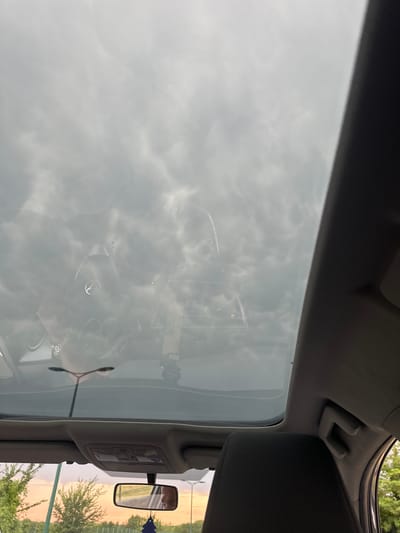 The image, taken from inside a vehicle, offers a view through the sunroof towards a cloudy sky and a reflection of the vehicle's interior and surroundings. The sky is overcast with dense, grey clouds. Reflected on the sunroof is the top of the car, including a circular emblem, likely a car manufacturer's logo, and part of the car's interior. Outside the car, trees and vegetation are visible, with a hint of an orange sunset in the distance. A car's rearview mirror hangs from the ceiling of the car, reflecting a person's profile and what appears to be a road. A blue car air freshener is also visible hanging from the mirror. The overall impression is of a cloudy day, possibly late afternoon or early evening, with the car traveling through a semi-urban or rural area. The location is identified as Tillé, France.