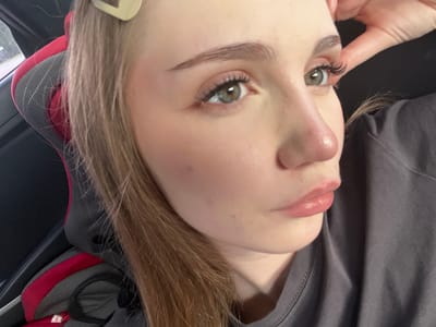 The image is a close-up, waist-up shot of a young woman with blonde hair, light skin, and green eyes. She is wearing a gray t-shirt and has false eyelashes. Her right hand is raised to her head. The background shows a car seat with red and gray accents. There is no indication of the city of Новая Отрадовка, Russia, in the image.