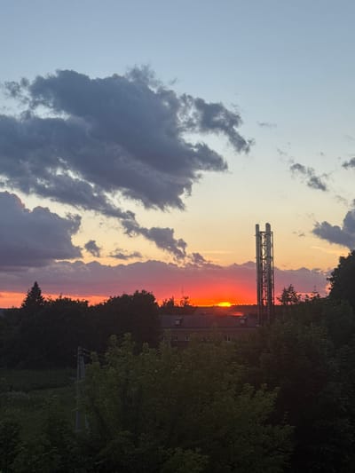 The image captures a sunset over Tovarkovo, Russia, with a vibrant orange sun low on the horizon. Dark, heavy clouds dominate the upper sky, contrasting with the clear, pale blue sky above. A large, industrial chimney structure stands prominently in the middle ground, silhouetted against the colorful sunset. The foreground is filled with lush, dark green trees and foliage, obscuring much of the ground below. A building with a pitched roof and multiple windows is partially visible through the trees, indicating a residential or industrial area. A utility pole with electrical wires is also visible on the left side of the frame.