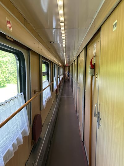 A photo taken from inside a train compartment. On the left side of the image are a series of windows with white, lace curtains. The walls are a light tan color with metal trim. The ceiling is a light-colored wood paneling, featuring a red emergency pull, silver metal handles, and an illuminated sign. The right side of the compartment has a dark brown padded backrest. At the far end of the compartment, a person is visible sitting down. The location is Радашковіцкі сельскі Савет, Belarus, as indicated by the metadata.