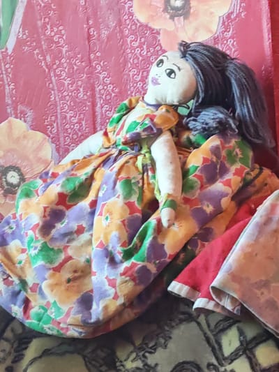 A close-up, slightly blurred shot features a handmade doll positioned in what appears to be a domestic setting. The doll, with a fabric head, painted eyes and lips, and dark yarn hair, is dressed in a brightly colored, floral-patterned dress. A portion of a red garment is visible to the right of the doll. The background consists of a textured pink surface with a floral motif and an adjacent area that is patterned with a dark, possibly embroidered, fabric. The overall impression is of a well-loved toy in a home environment, potentially in Чонкаўскі сельскі Савет, Belarus, though this location is not visually confirmed.