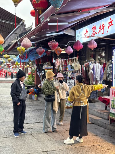The image depicts an outdoor market scene in Pengshui Miao and Tujia Autonomous County, China, under an overcast sky. The street is paved with grey rectangular stone slabs.
In the foreground, four individuals are visible. From left to right:
1. A male individual wearing a black knit beanie, a black jacket over a light pink collared shirt, black trousers, and blue sneakers. He carries a dark grey patterned crossbody bag and holds a small orange object in his right hand. He is facing to his right.
2. A female individual wearing a light brown bucket hat, an olive-green jacket, light blue wide-leg jeans, and brown boots. She carries a dark crossbody bag and a light beige fringed scarf. Her head is tilted downwards, and she appears to be looking at a smartphone held in her hands. She is positioned slightly to the right of the male.
3. A female individual wearing a light pink bucket hat, a light beige fluffy jacket, brown trousers, and white sneakers. She is standing next to the second female, facing towards her left, with her mouth slightly open, suggesting speech or a reaction.
4. A female individual, seen from behind, wearing a mustard yellow jacket with white ties (possibly an apron), a black wide-leg skirt or trousers, and white chunky sneakers. Her dark hair is pulled back. She wears yellow sleeves or gloves on her forearms. Her right arm is extended horizontally to the right, in a pointing or gesturing motion. She is positioned facing away from the viewer, addressing the group in front of her.
The background features numerous vibrant, decorative paper lanterns of various shapes and colors (red, pink, orange, yellow, green, blue) suspended across the street above market stalls. Two stalls are visible with dark red awnings. The stall on the right has a prominent illuminated sign displaying red Chinese characters, "丸黎特产" (Wán Lí Tèchǎn), which translates to "Wanli Specialty Products." This stall displays clothing items, including knitted sweaters and scarves, along with bags and other merchandise. A table in the lower right foreground displays a white and green sign with the Chinese character "鲜" (Xiān, meaning fresh). Traditional Chinese architectural elements with dark, curved roofs are partially visible behind the stalls. A blue umbrella is visible under some lanterns near the left stall. Green potted plants are also discernible near the left side of the market area. Further down the street, blurred figures suggest more pedestrian traffic.