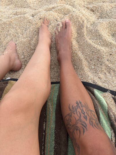 The image shows a close-up, high-angle view of two people's legs and feet resting on a sandy beach, with a striped towel underneath. The legs are positioned diagonally, with the left leg appearing closer to the camera and the right leg further away. The sand is light brown and speckled with small, dark particles. The left leg is lighter in skin tone and appears smooth, with some sand grains visible on the foot. The right leg is darker in skin tone and has a large tribal tattoo on the lower calf. The foot of the right leg is also visible, with toes slightly separated. The towel has horizontal stripes in shades of brown, green, and black. The scene appears to be a casual, outdoor setting at a beach. No specific landmarks in Vilassar de Mar, Spain are identifiable.