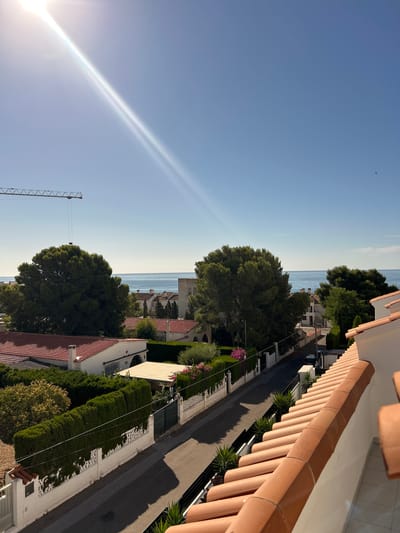 The image captures a sunlit residential street scene in Alcalà de Xivert, Spain, with a view towards the Mediterranean Sea. A bright sunbeam cuts diagonally across a clear blue sky. In the foreground, terracotta roof tiles are visible on a balcony railing. The street, lined with white fences and manicured hedges, curves to the right. Several houses with red tile roofs are nestled amongst mature green trees. Beyond the houses, the blue expanse of the sea is visible under a cloudless sky. A construction crane is positioned in the upper left quadrant of the image. A small dark object, possibly a bird, is visible in the far upper right of the sky. No people are depicted in the image.
