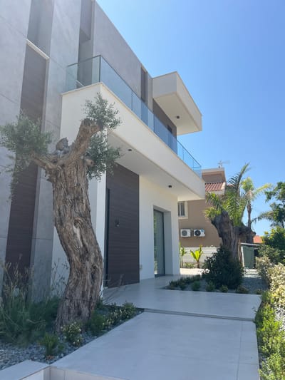 A modern house with a striking architectural design is shown in an exterior shot. The house features clean lines, a mix of concrete and wood-paneled facades, and large glass windows and balconies. A prominent, gnarled olive tree trunk extends horizontally across the foreground, its rough bark and sparse foliage creating a dramatic contrast with the sleek building. The setting is a residential area, with glimpses of neighboring houses and lush greenery. The sky is clear and blue, indicating a sunny day. Two outdoor air conditioning units are visible mounted on the beige wall of the house. The scene appears to be set in Moni Community, Cyprus. There is no visible text in the image.