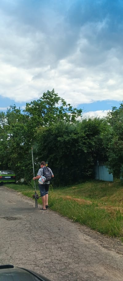 A full-shot, eye-level, vertical, outdoor, daylight, slightly overcast image shows a person walking away from the camera on a cracked asphalt road. The person is wearing a striped t-shirt, blue shorts, and flip-flops, with a backpack on their shoulders. They are carrying a fishing rod, a white bucket with red and blue markings, and what appears to be two plastic bottles or glasses on their back. To the left of the road, a green car is parked. The background is filled with dense green trees and bushes. The sky above is a mix of blue and white clouds. The location is identified as Popivka, Ukraine.