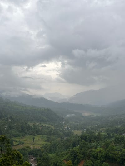 The image depicts a hilly landscape in Ramboda, Sri Lanka, under overcast skies. Dense vegetation covers the hillsides, with hints of rice paddies and small settlements scattered throughout. Mountains fade into the distance, obscured by mist and low clouds. The sky is dominated by gray clouds, with a patch of brighter light breaking through.