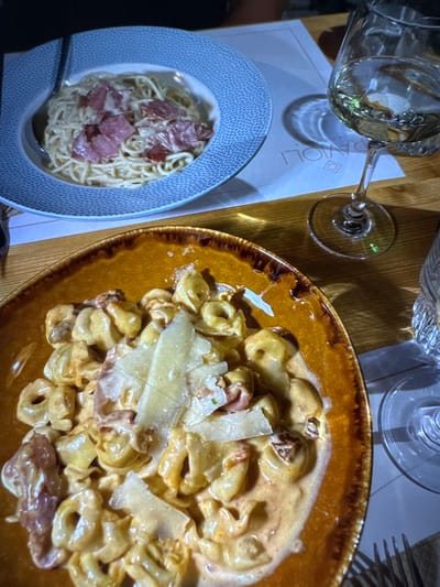 The image displays a close-up, overhead view of two pasta dishes and accompanying drinks on a wooden dining table. The scene is located at Ravioli Restaurant in Kumanovo, North Macedonia, as indicated by the "RAVIOLI" text visible on a white placemat or menu beneath a wine glass.

In the upper left, a light blue bowl with a stippled rim contains spaghetti coated in a light-colored sauce, topped with several irregular, reddish-purple pieces of cured meat. To the lower right, a rustic mustard-yellow and brown bowl holds tortellini or similar stuffed pasta in a rich, creamy orange-hued sauce, garnished with multiple shavings of pale yellow Parmesan cheese and smaller fragments of cured meat.

Adjacent to the upper pasta dish and central to the right side of the frame, a stemmed wine glass holds a clear, pale yellow liquid, consistent with white wine. Its base rests on the aforementioned white paper item. A portion of another clear drinking glass and a silver fork are visible in the bottom right corner. No individuals are visible in the frame; the scene depicts a prepared meal setting.
