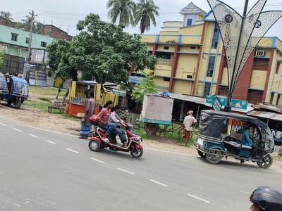 The image depicts a street scene in Benapole, Bangladesh. In the foreground, a red three-wheeled electric scooter with two occupants is moving along the road. The rider is wearing a light-colored shirt and jeans, while the passenger behind him is wearing a pink shirt and jeans. A blue electric rickshaw with a driver and a passenger is visible on the right side of the road. The rickshaw is black and blue and has a canopy. Further down the road, a man in a white shirt and a tan lungi is standing next to a bicycle. Behind the road, there are several shops with awnings and merchandise displayed. A large green tree with dense foliage is prominent in the mid-ground. Several buildings are visible in the background, including a yellow building with blue trim and a light green building with a dark roof. Palm trees are also present in the background, along with utility poles and overhead wires. The sky is overcast.