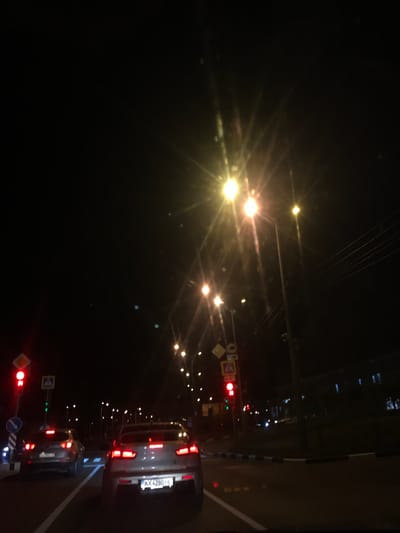 The image depicts a nighttime street scene. The perspective is from inside a vehicle looking forward. In the foreground, the rear of a silver sedan is visible, displaying its red taillights and a license plate reading "AX 4280 HL". To the left, another vehicle, a silver SUV, is also seen from the rear, its taillights illuminated.
The street is illuminated by multiple streetlights, some of which create starburst effects due to the camera's aperture. Traffic signals are present; a red traffic light is clearly visible on the left, alongside pedestrian crossing and other traffic signs. The road surface appears to be asphalt, with white lane markings. In the background, buildings and more streetlights are faintly discernible in the darkness. The overall environment suggests an urban setting at night, with typical traffic and street infrastructure.