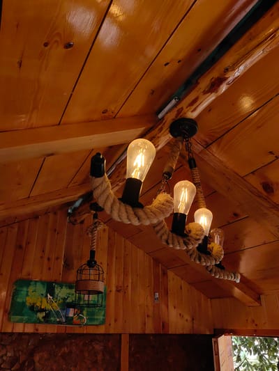 The image shows a rustic chandelier made of thick rope, hanging from a wooden ceiling. The chandelier has four Edison-style light bulbs, which are lit. To the left of the chandelier, there is a decorative wall hanging with abstract green and yellow patterns. The walls and ceiling are made of polished wood planks. The scene appears to be indoors, possibly in a cabin or a restaurant, given the wooden decor. There are no people visible in the image. The overall impression is one of a warm, cozy, and somewhat rustic ambiance. The image does not contain information about the city of Chhyal, India.