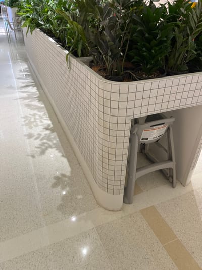 The image, rotated ninety degrees counter-clockwise, captures an indoor public environment, likely a shopping center in Indooroopilly, Australia. The central subject is a large, white planter box or raised garden bed, featuring a grid-like pattern that resembles small tiles or a printed overlay. The planter's edge is smoothly curved on the right side.

Within the planter, dense, dark green foliage of robust indoor plants, possibly a species like Zamioculcas zamiifolia (ZZ plant), spills over the edges. The plants appear healthy, with some visible roots near the soil surface.

Behind the planter, tucked into a recessed area, is a gray, foldable baby changing station or a high chair, stored in an upright, folded position. A white warning label with indecipherable black text is visible on its side, likely containing safety instructions or usage information.

The floor is a light-colored terrazzo or speckled material, accented with subtle, lighter beige striped patterns. Overhead lighting casts distinct, leaf-shaped shadows from the plants onto the reflective floor surface, indicating a brightly lit space. No people are present, and no activity is taking place, presenting a static scene of a well-maintained modern interior.