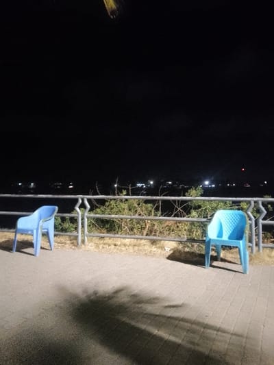 Two light blue plastic chairs sit on a paved promenade at night. A metal railing runs along the edge, with sparse greenery growing behind it. Across a dark expanse of water, a faint line of lights indicates a distant shore. The sky is a vast black void. Shadows from unseen objects stretch across the pavement towards the viewer, adding a layer of mystery to the scene.