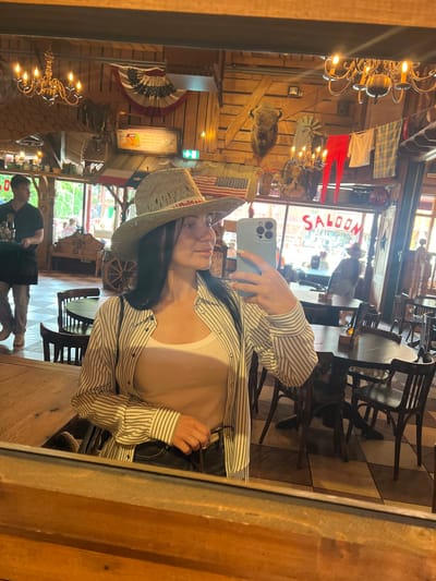 The image depicts a reflection in a mirror of a woman in a straw cowboy hat, a striped button-up shirt, and a white tank top, taking a selfie with her phone. She is seated at a wooden table in a restaurant with a Western theme, potentially the Saloon restaurant in Collendoorn, Netherlands, based on the visible "SALOON" signage. The background of the reflection reveals the restaurant's interior, which features wooden paneling, chandeliers, mounted animal heads, and American flags. Several tables and chairs are visible, as are other patrons. The woman's hands are visible, one holding the phone and the other resting on her lap, adjusting her belt. The overall scene suggests a casual dining or entertainment environment with a distinct Wild West aesthetic.