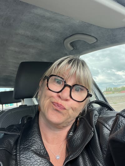 A woman with blond hair and black-rimmed glasses is taking a selfie while seated in a car. She is wearing a black leather jacket and a silver necklace with a circular pendant. The interior of the car features a gray headliner with a grab handle. Outside the car window, there is a glimpse of a building and trees, suggesting the location is in Saint-Cyprien, France.